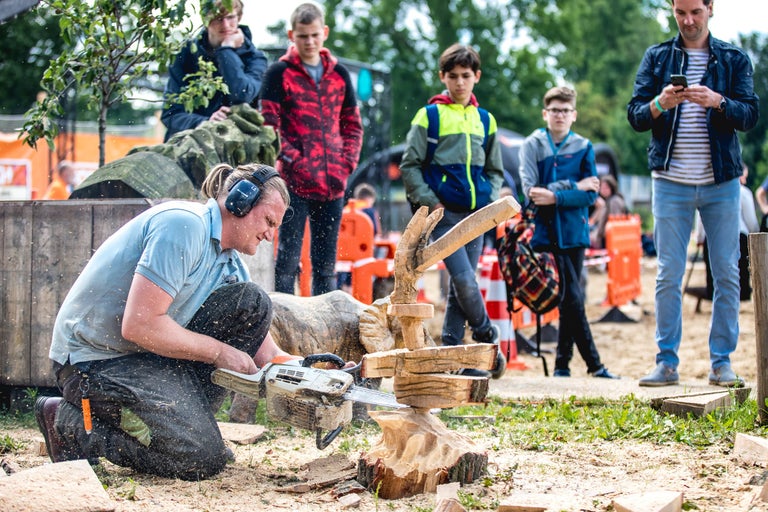 Een man knielt en werkt met een kettingzaag aan een houten sculptuur, omringd door toeschouwers.