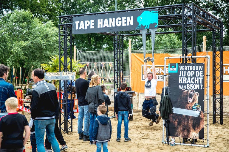 Scene met persoon die aan een hindernisbaan hangt, met de tekst Tape Hangen en Bison logo