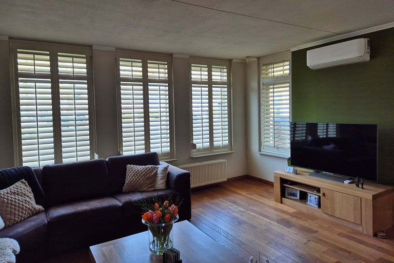 Woonkamer met ramen voorzien van lichte luiken, donkere leren bank, houten tafel, houten vloer en televisie op een houten tv-meubel.