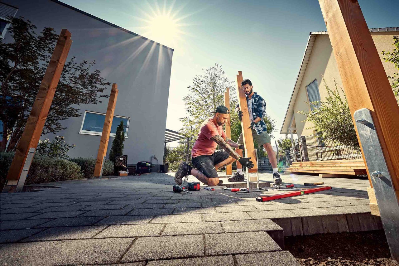 Twee ambachtslieden bouwen een houten constructie in de tuin met gereedschap zoals een boormachine, waterpas en lijmklemmen.