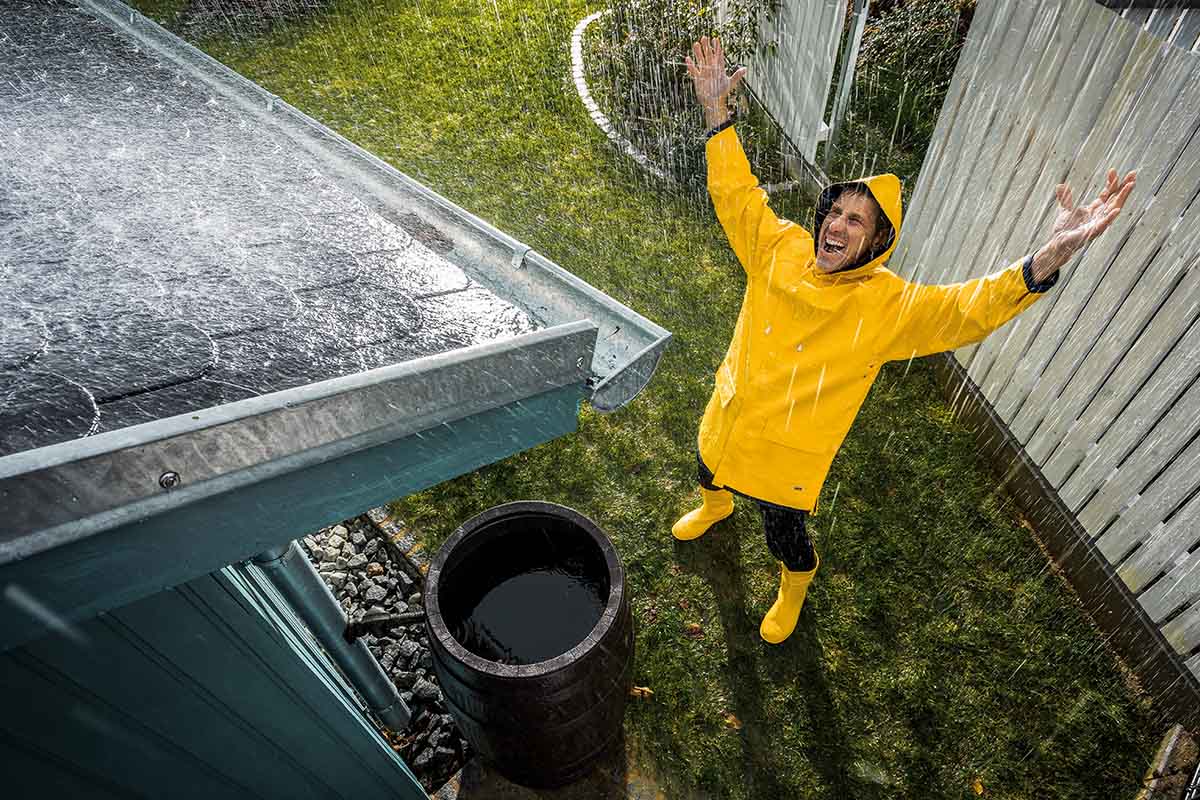 Man in regenkleding staat in de zware regen naast een regenton die onder een dakgoot is geplaatst voor het opvangen van regenwater.