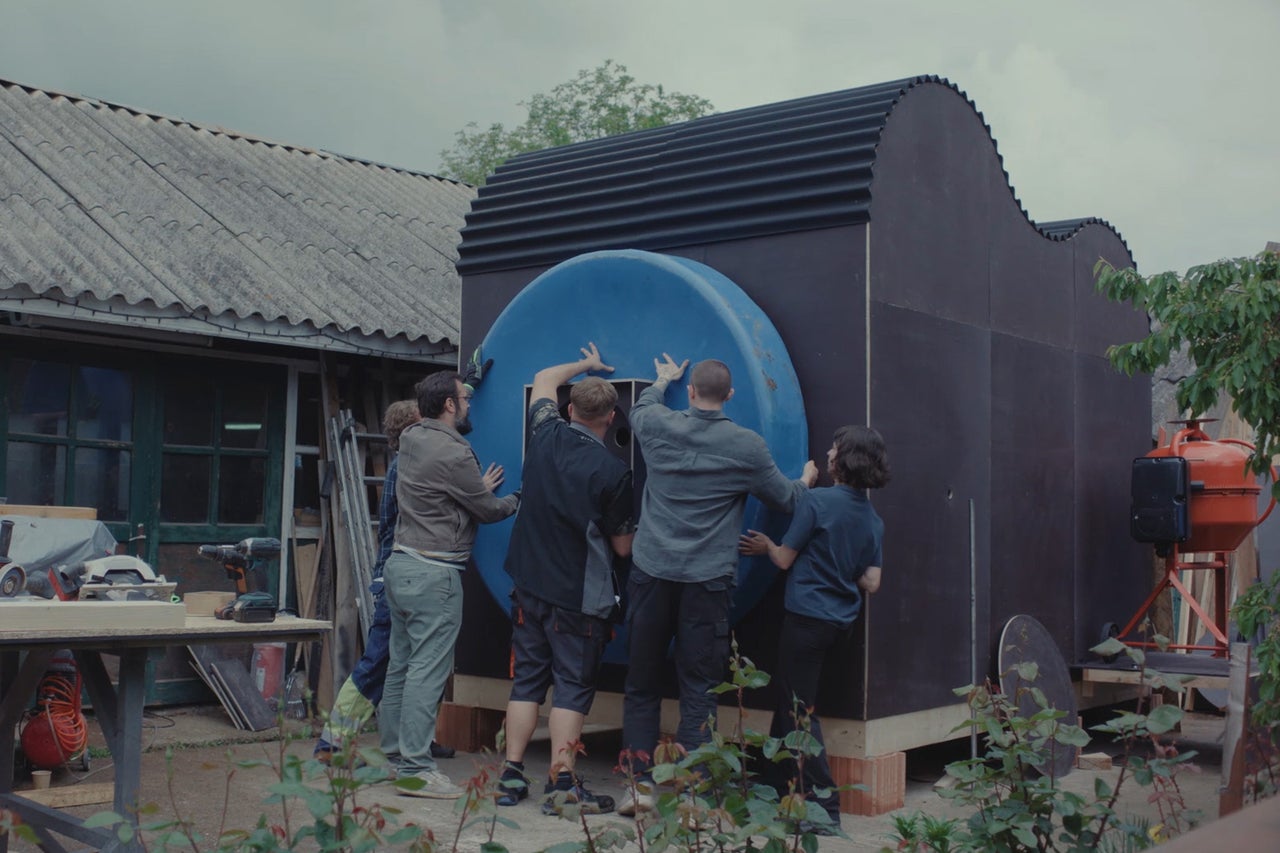 Een groep mensen werkt samen aan de bouw van een moderne zwarte tuinkamer met een groot rond blauw element.