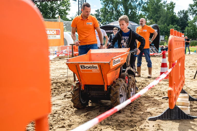 Scene met een kind dat een minidumper bestuurt, omringd door volwassenen en afzettingen op een zandachtige ondergrond