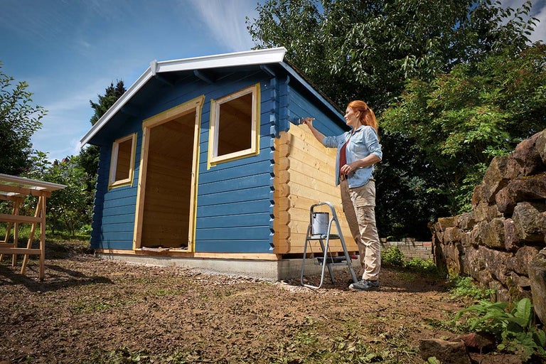Een vrouw schildert een houten tuinhuis met blauwe verf.
