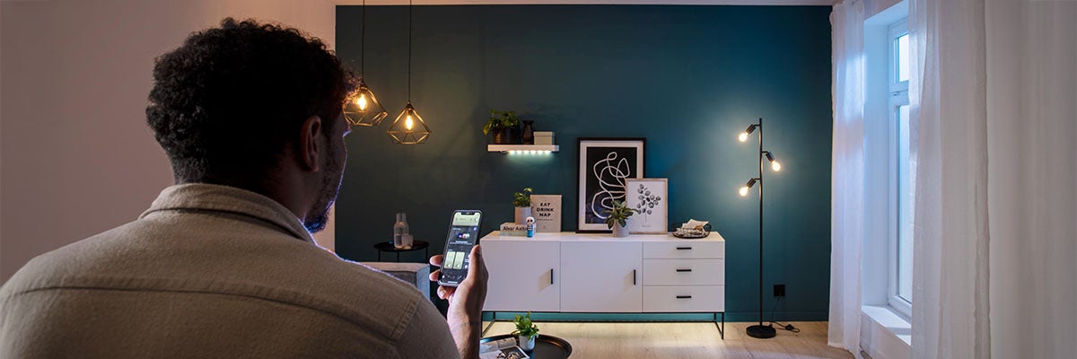 Persoon bedient slimme verlichting met een app in een modern ingerichte woonkamer met dressoir en wandlampen.