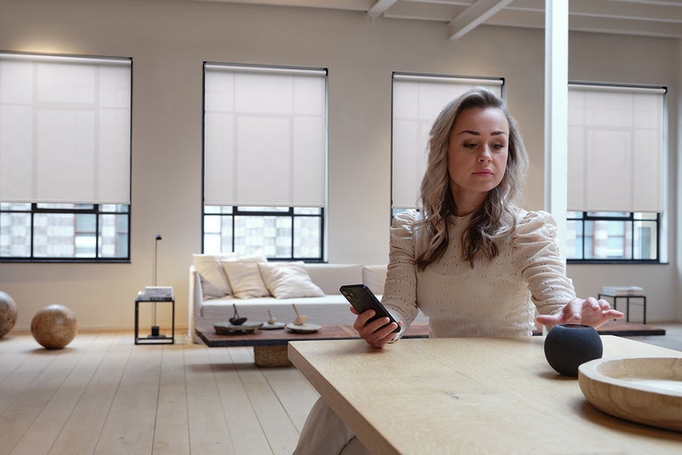 Een vrouw in een moderne, lichte woonkamer bedient via smartphone en smart speaker vier automatische rolgordijnen voor grote ramen.