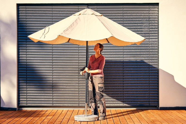 Vrouw in werkkleding staat op een houten terras naast een grote beige parasol met een donkere mast voor een grijze rolluikwand