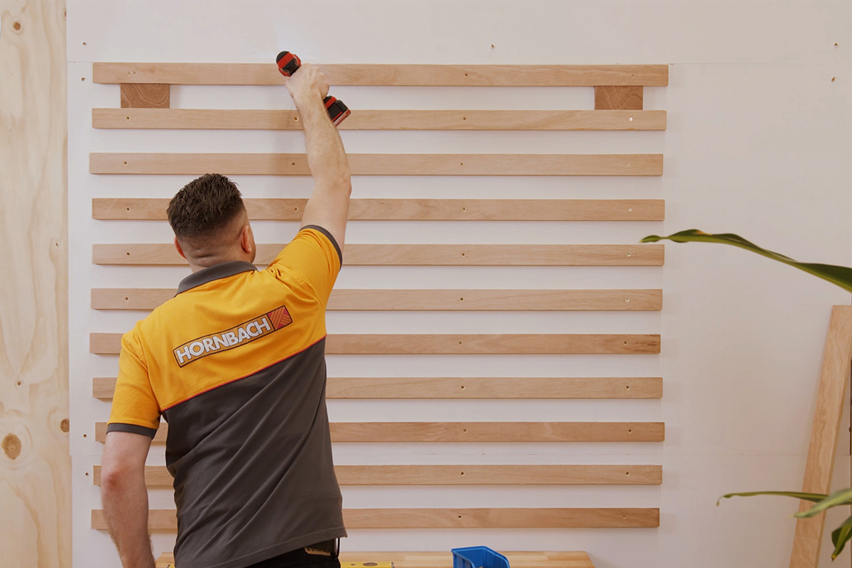 Vakman bevestigt houten latten met een accuschroevendraaier aan een muur. Hornbach Logo.