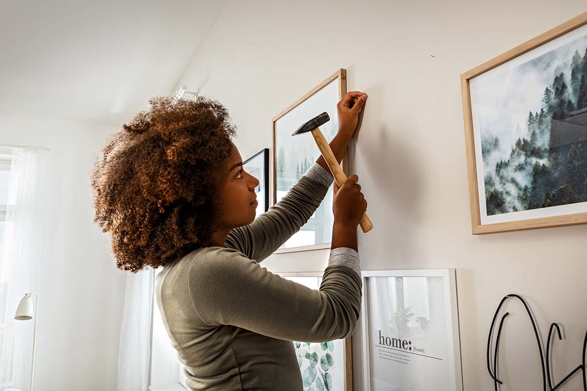 Een vrouw slaat met een hamer een spijker in een muur om verschillende fotolijsten op te hangen in een lichte kamer.