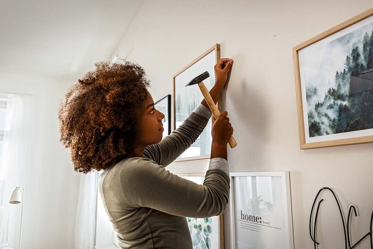 Een vrouw slaat met een hamer een spijker in een muur om verschillende fotolijsten op te hangen in een lichte kamer.