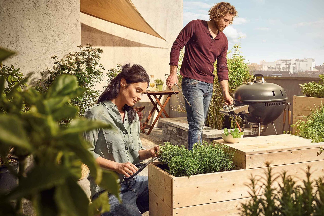 Vrouw snoeit kruiden in een verhoogde tuinbak, man grilt op de achtergrond op het balkon