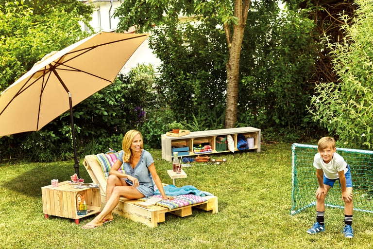 Tuinlandschap met vrouw op houten pallet onder parasol en spelend kind bij voetbaldoel