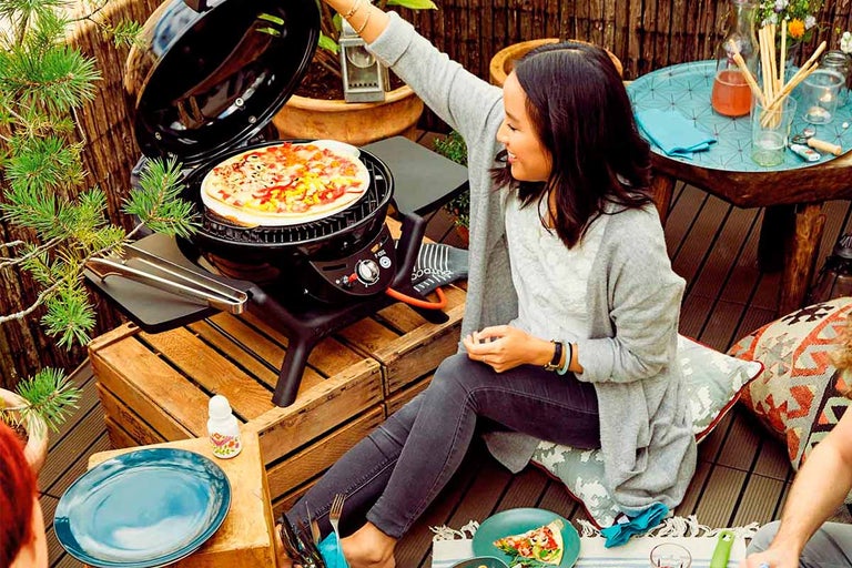 Scène met een vrouw zittend naast een barbecue met pizza, omringd door borden, drankjes en decoraties voor een ontspannen maaltijd buitenshuis.