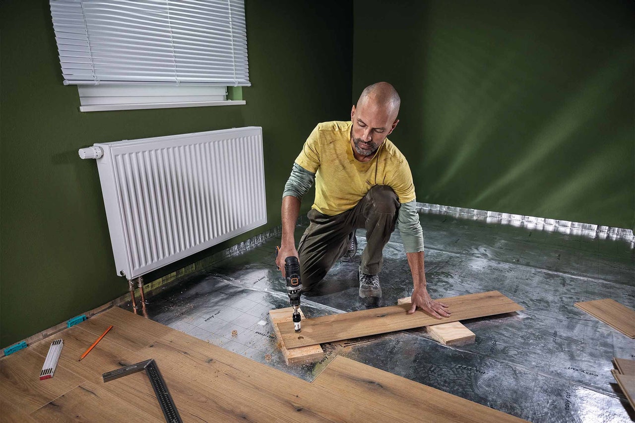 Een man legt laminaat met een boormachine in een kamer met een radiator en jaloezieën.