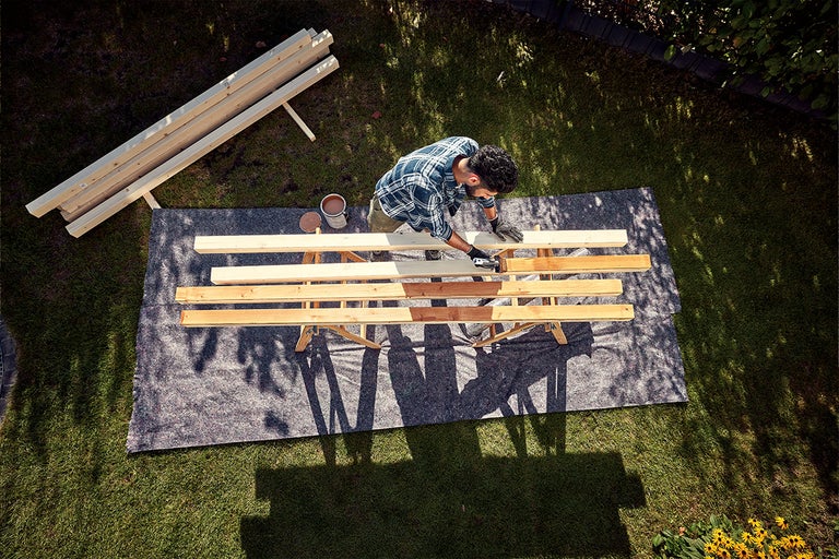 Man schildert houten planken in de tuin met een kwast, naast hem hout en een verfblik.