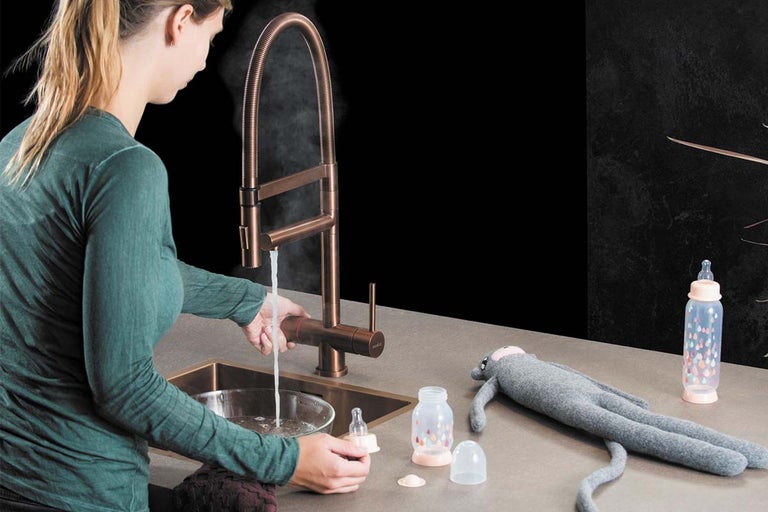 Een persoon gebruikt een koperkleurige professionele keukenkraan met flexibele spiraalveer om babyaccessoires af te spoelen met stomend water.