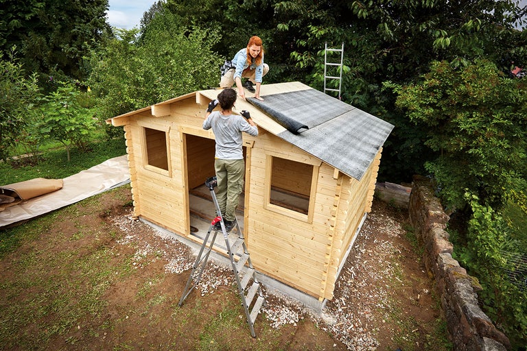 Twee mensen monteren dakleer op een houten tuinhuis.