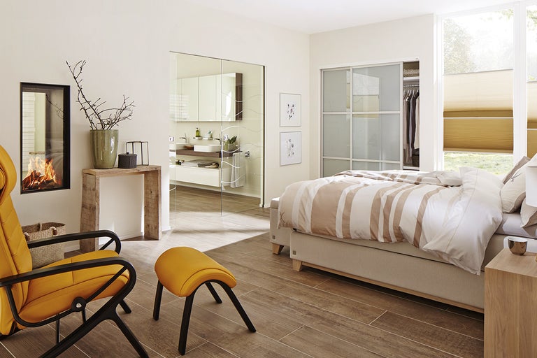 Lichte slaapkamer met open haard, bed, fauteuil, voetenbank en toegang tot de badkamer via een glazen schuifdeur.