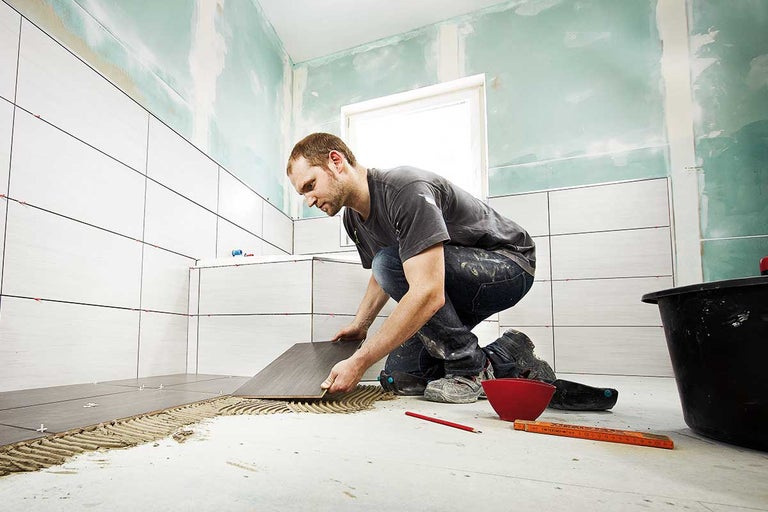 Een tegelzetter legt tegels in de badkamer. Er zijn gereedschappen en bouwmaterialen in de ruimte te zien.