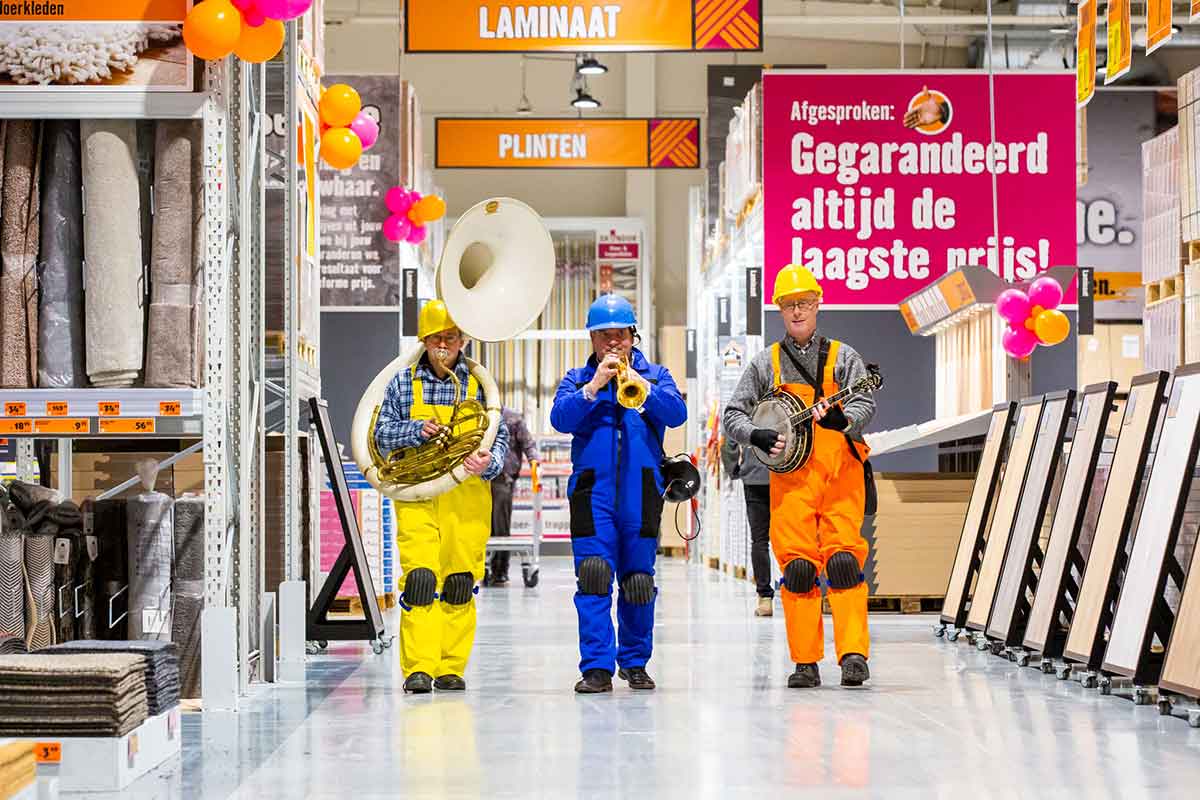 Drie muzikanten met tuba, trompet en banjo in een Hornbach vestiging tussen tapijten, laminaat en plinten.