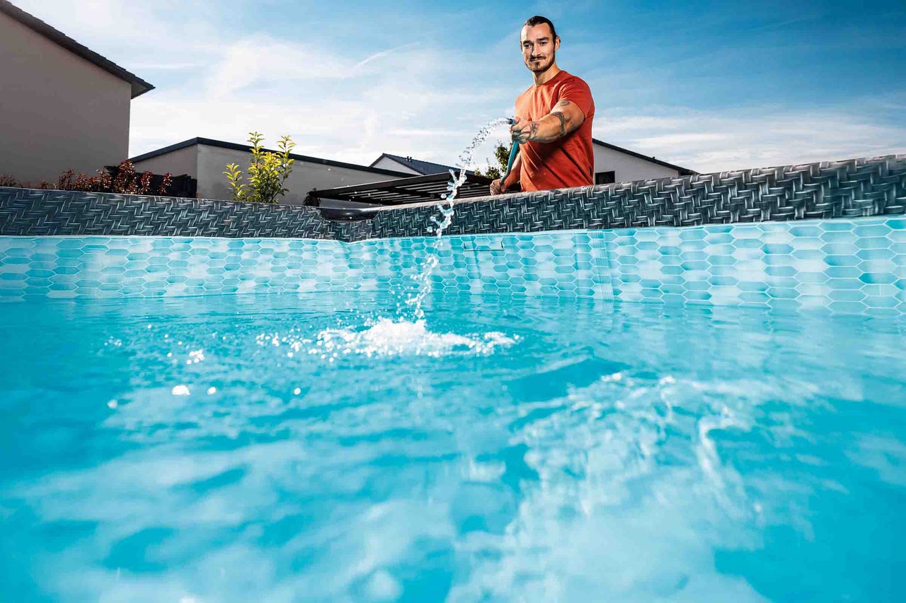 Een man vult een bovengronds zwembad met rotan-look en mozaïekprint met water uit een tuinslang.