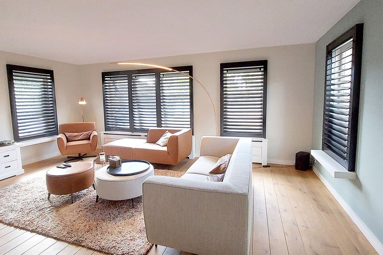 Woonkamer met houten vloer, banken, fauteuils, tapijt en houten jaloezieën voor lichtregulering