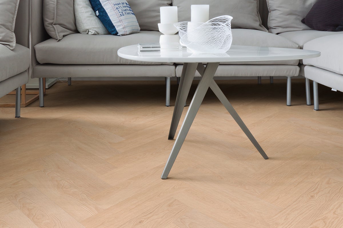 Lichte laminaatvloer met visgraatmotief in een moderne woonkamer met grijze bank en witte salontafel.