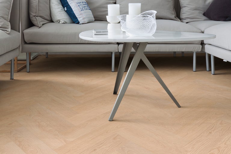 Lichte laminaatvloer met visgraatmotief in een moderne woonkamer met grijze bank en witte salontafel.