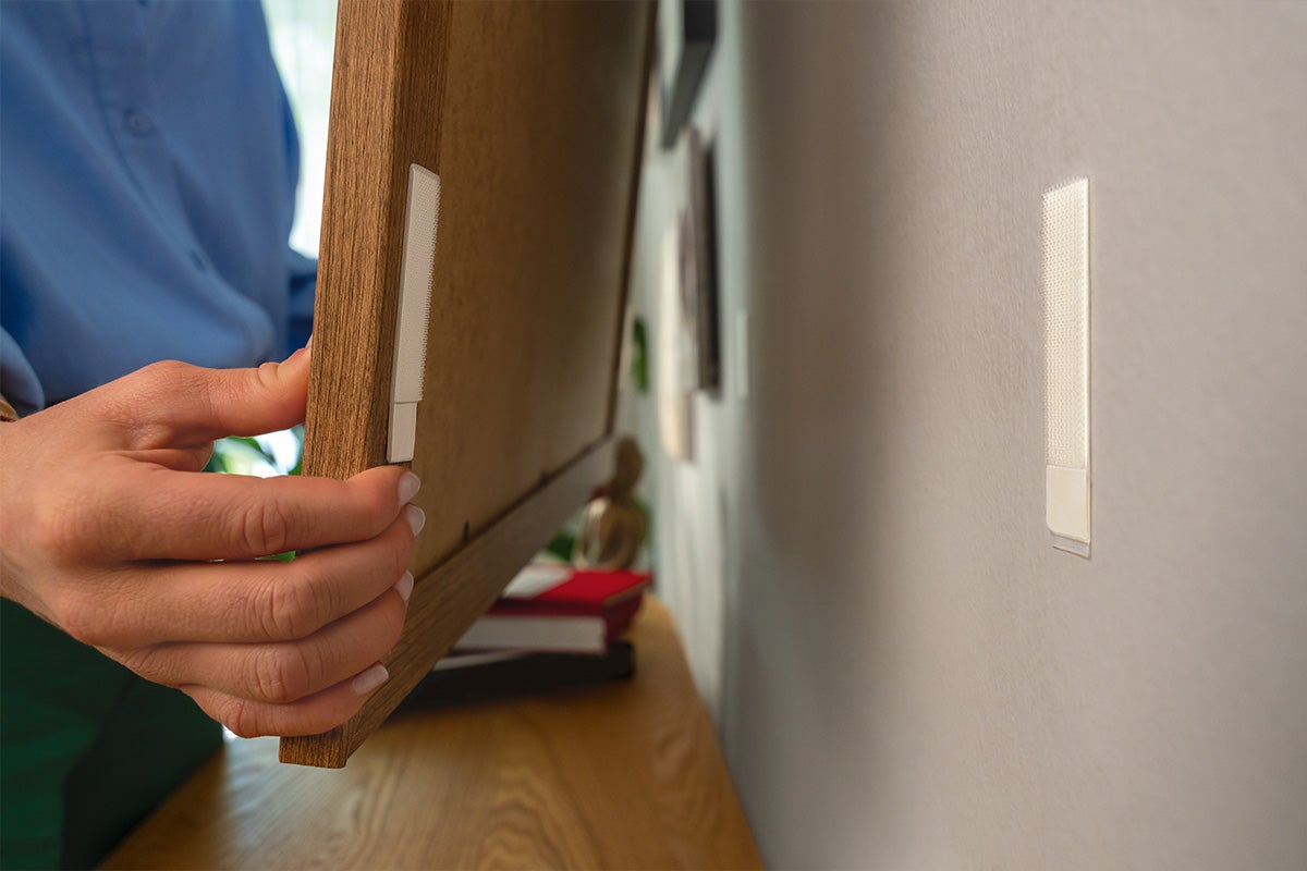 Iemand brengt zelfklevende strips aan op een houten lijst en een muur om schilderijen op te hangen zonder te boren.