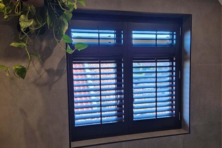 Moderne shutters met verstelbare lamellen voor een raam aan de binnenzijde, gedecoreerd met een hangplant aan een donkere muur.