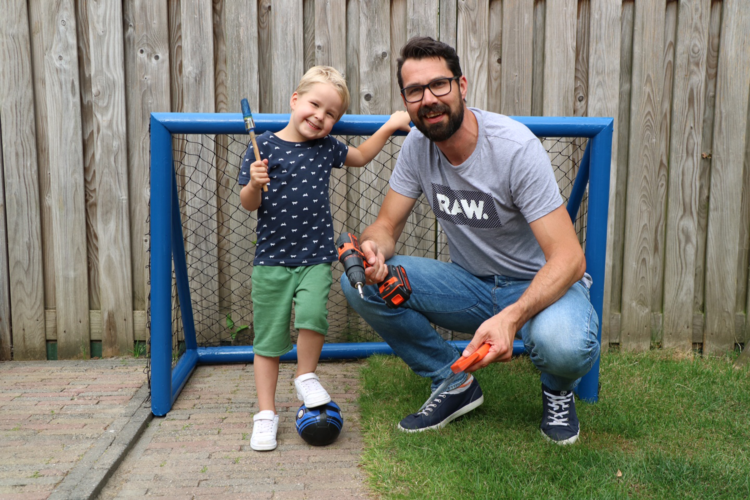 Vader en zoon staan met gereedschap en voetbalgoal in de tuin.