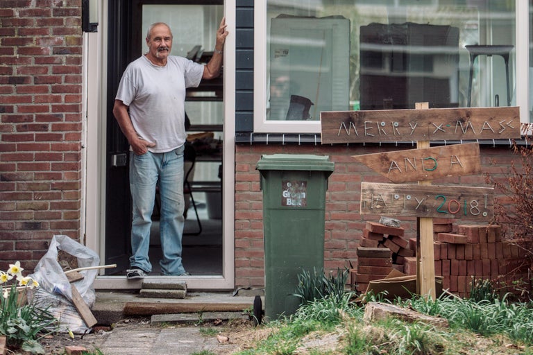 Man staat in de deuropening van een bakstenen huis met een afvalcontainer en tuindecoratie