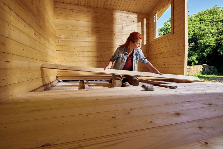 Vrouw legt houten vloer in een tuinhuis met hamer en schroevendraaier.