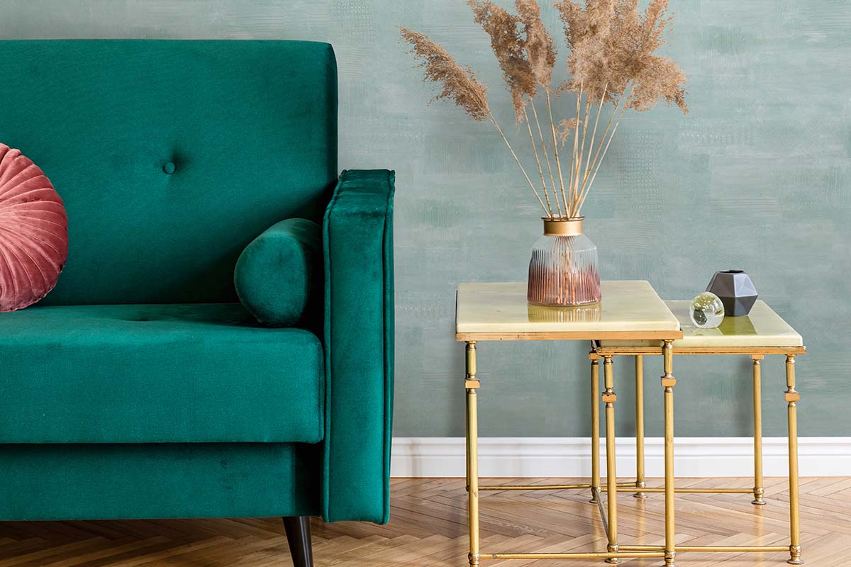 Woonkamer met een groene fluwelen bank, twee bijzettafels met metalen frame en een glazen vaas met droogbloemen tegen een gestructureerde wand.