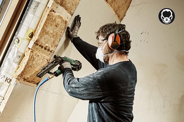 Ambachtsman bevestigt gipsplaat met een pneumatische tacker aan een dakhelling. Hij draagt een beschermend masker en gehoorbescherming.