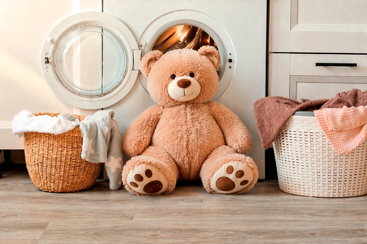 Een grote teddybeer zit voor een geopende wasmachine tussen twee wasmanden op een lichte vloer met houtlook.
