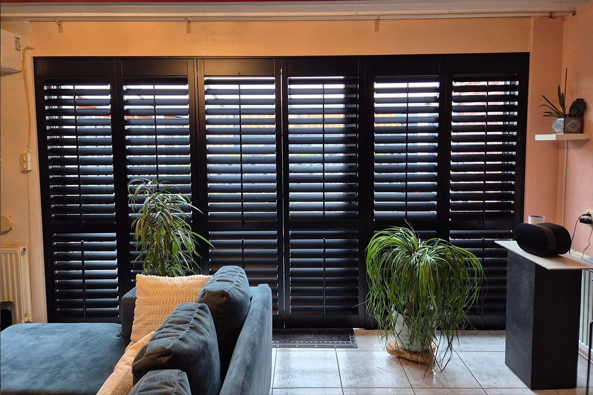 Woonkamer met kamerhoge ramen, voorzien van donkere shutters, gecombineerd met kamerplanten en een bank.