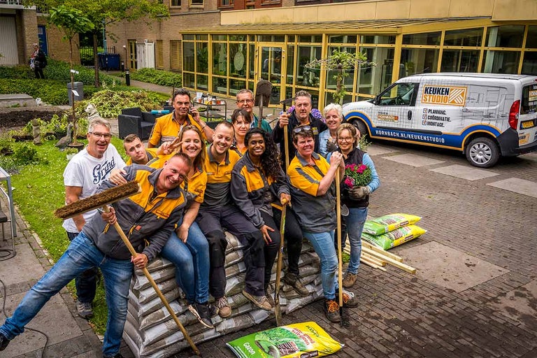 Een groep Hornbach-medewerkers poseert voor een Hornbach keukenstudio busje, tuingereedschap en zakken aarde.