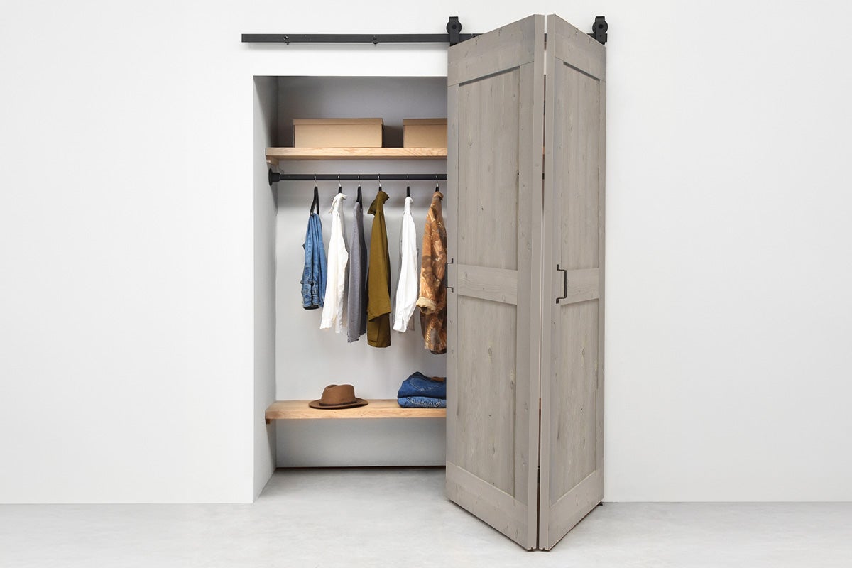 Vouwschuifdeur in barndoor-stijl van grijs hout aan een zwarte rail voor een kledingkast met kleding en planken.