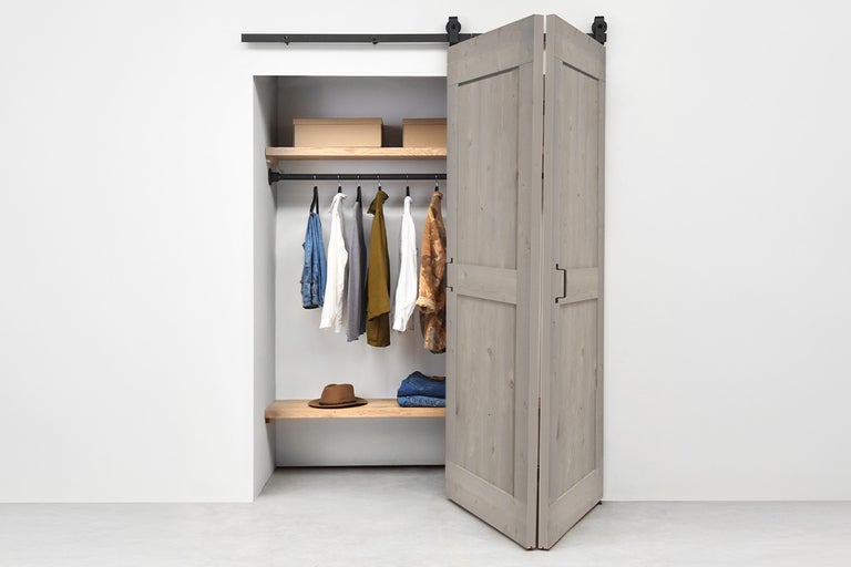 Vouwschuifdeur in barndoor-stijl van grijs hout aan een zwarte rail voor een kledingkast met kleding en planken.