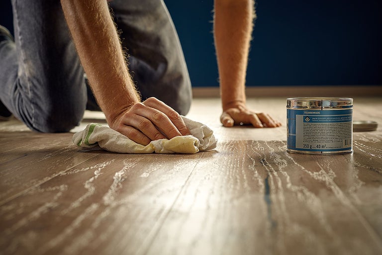 Een persoon brengt vloerwas aan op een houten vloer met een doek. Er staat een blik vloerwas naast.