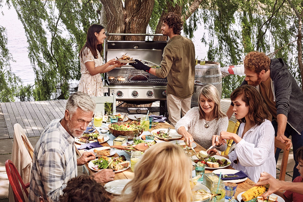 Een groep mensen geniet van een maaltijd in de buitenlucht met een barbecue op de achtergrond.