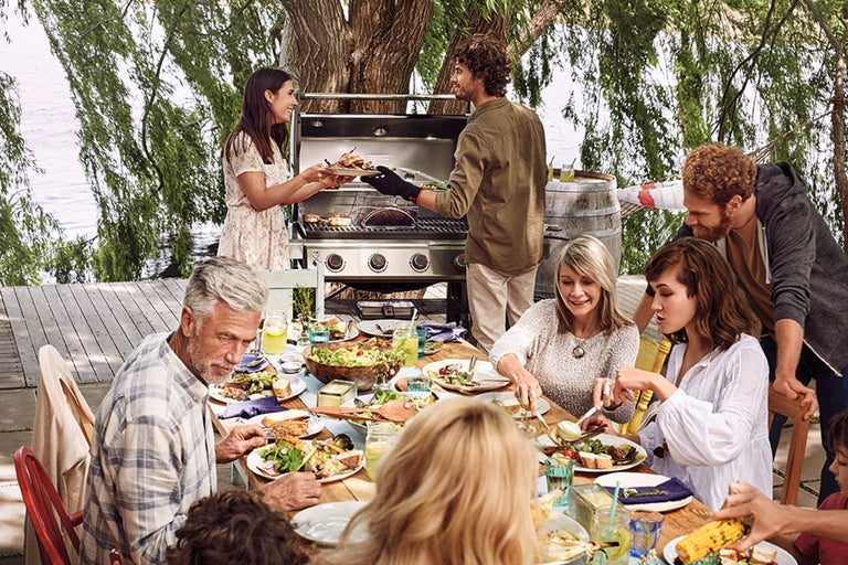 Een groep mensen geniet van een maaltijd in de buitenlucht met een barbecue op de achtergrond.