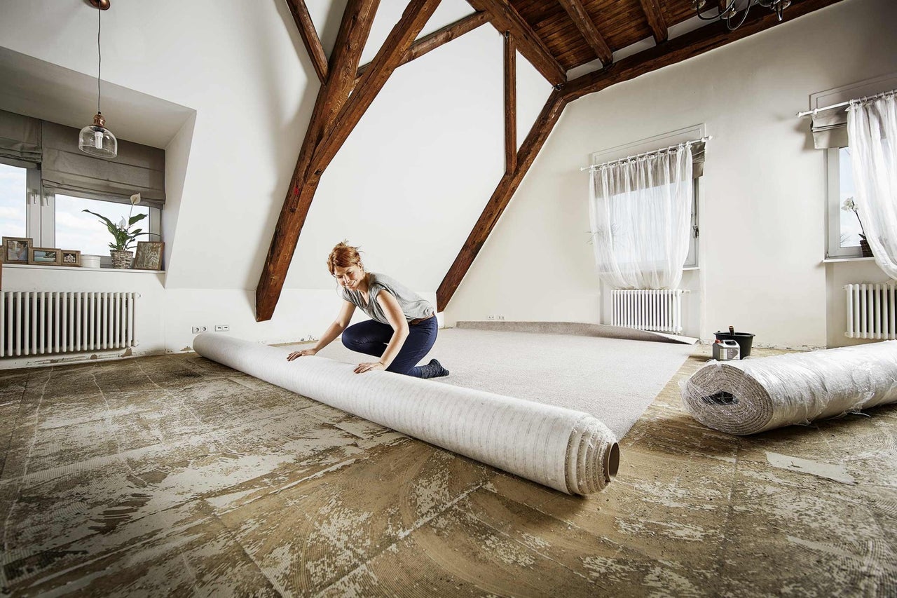 Vrouw rolt tapijt uit in een zolderkamer. Houten balken zijn zichtbaar.