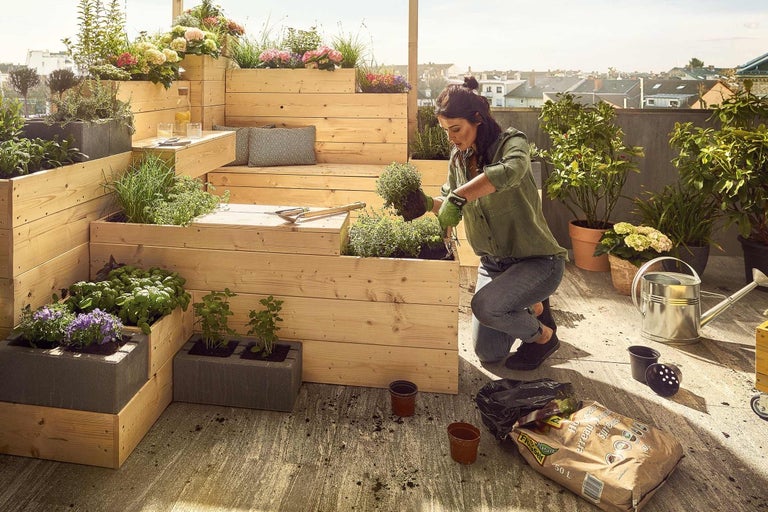 Een vrouw beplant een houten terras met meerdere niveaus met verschillende planten, kruiden en bloemen in plantenbakken en potten.