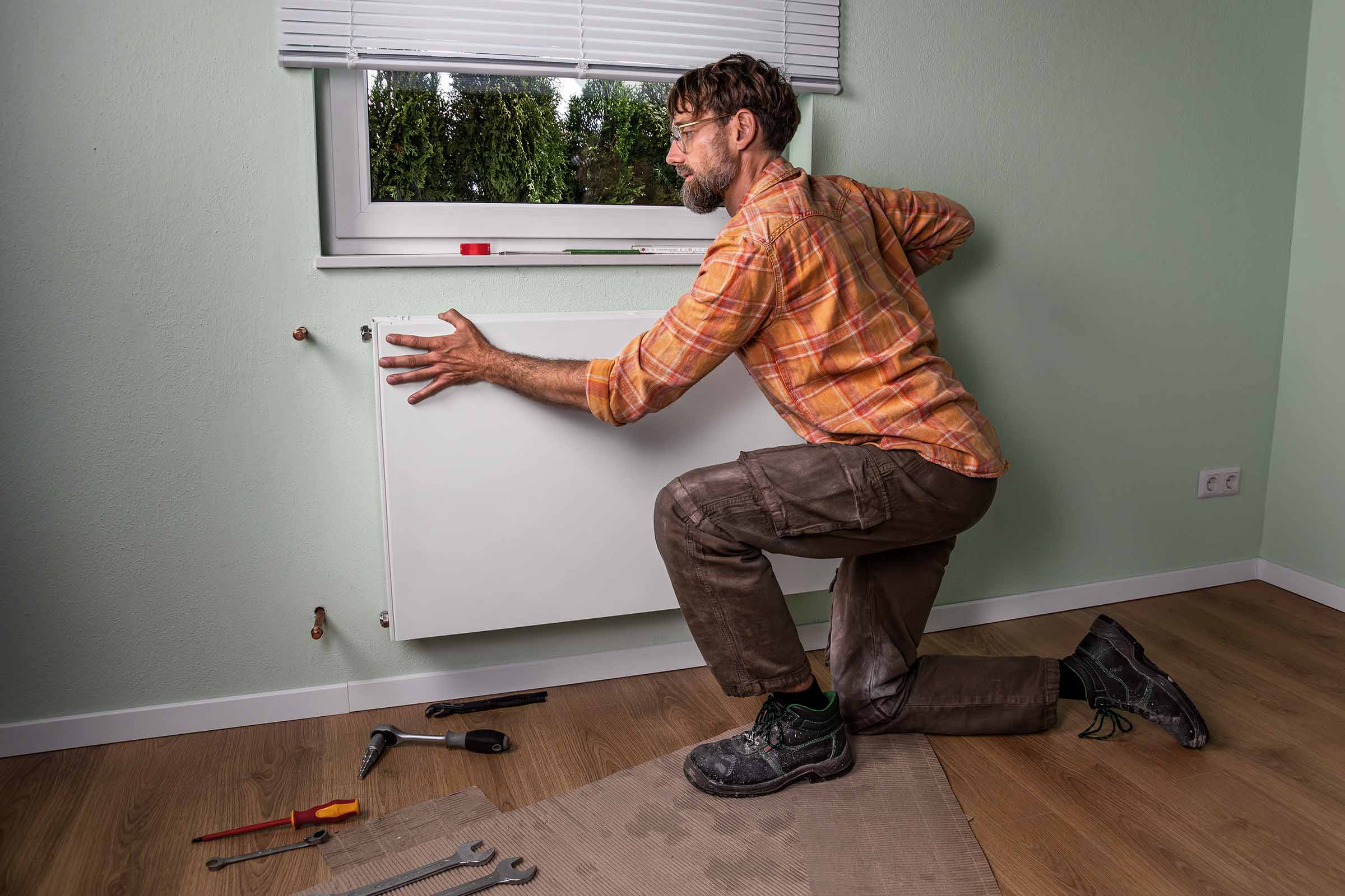 Een man knielt voor een muur en monteert een radiator. Er ligt gereedschap op de vloer.