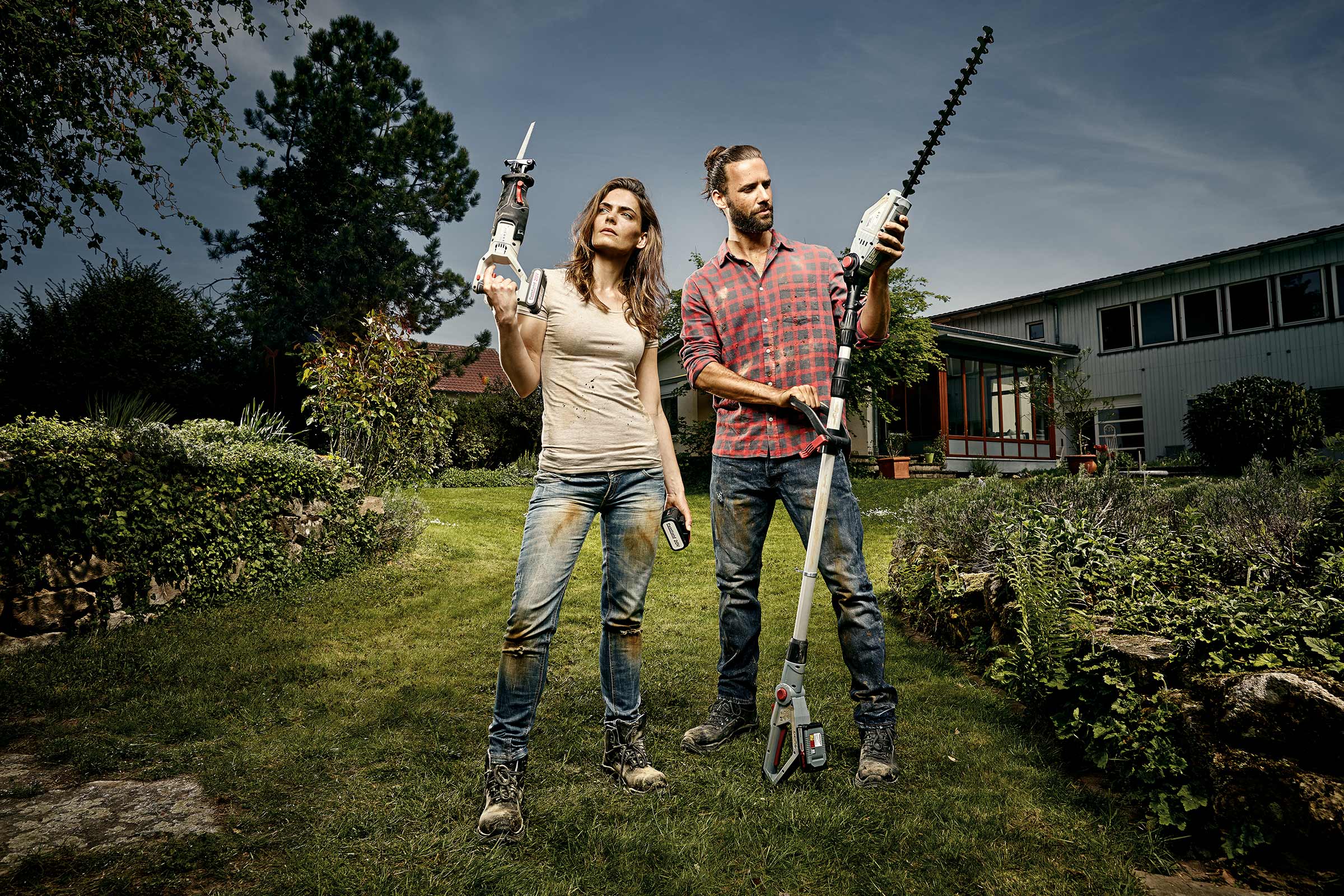 Een man en een vrouw staan met een heggenschaar en een reciprozaag in een tuin.