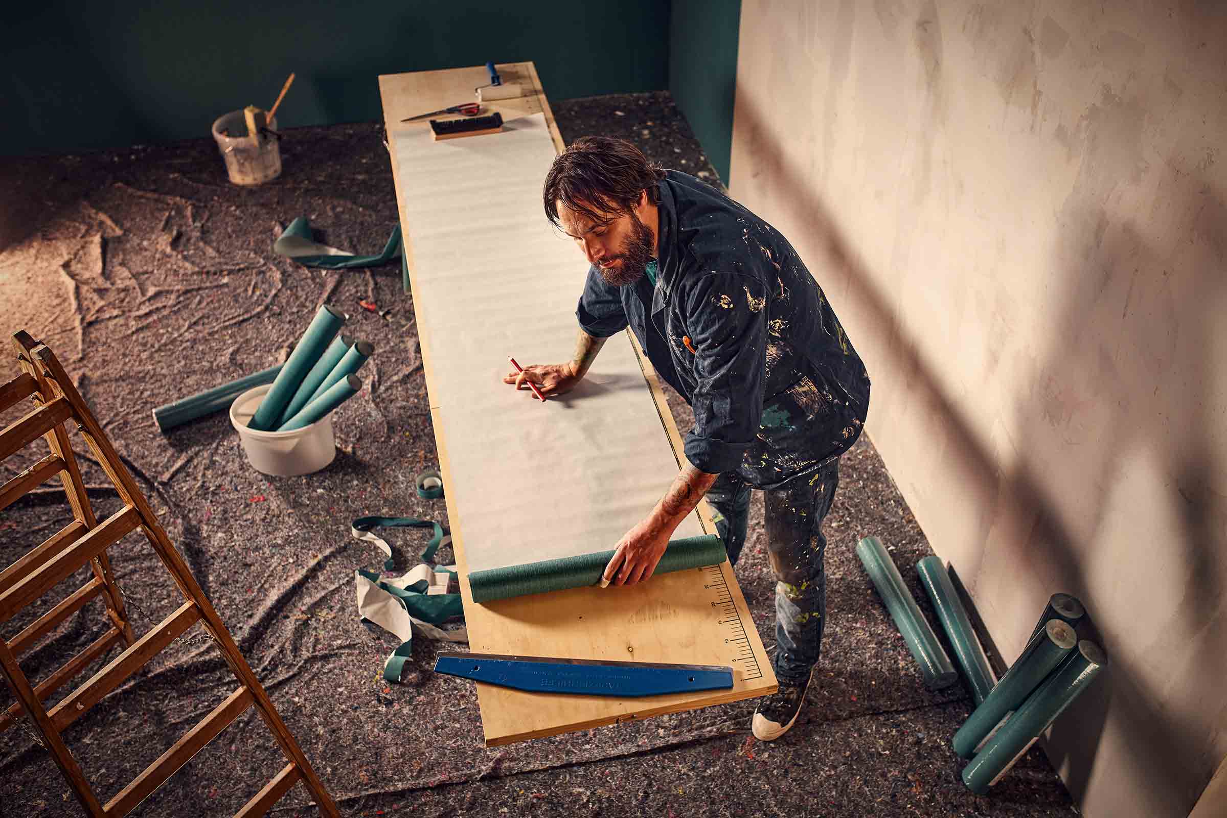 Een man bereidt een behang voor op een tafel om het aan de muur aan te brengen. Gereedschappen zoals een ladder, een emmer en een behangroller zijn verspreid in de kamer.