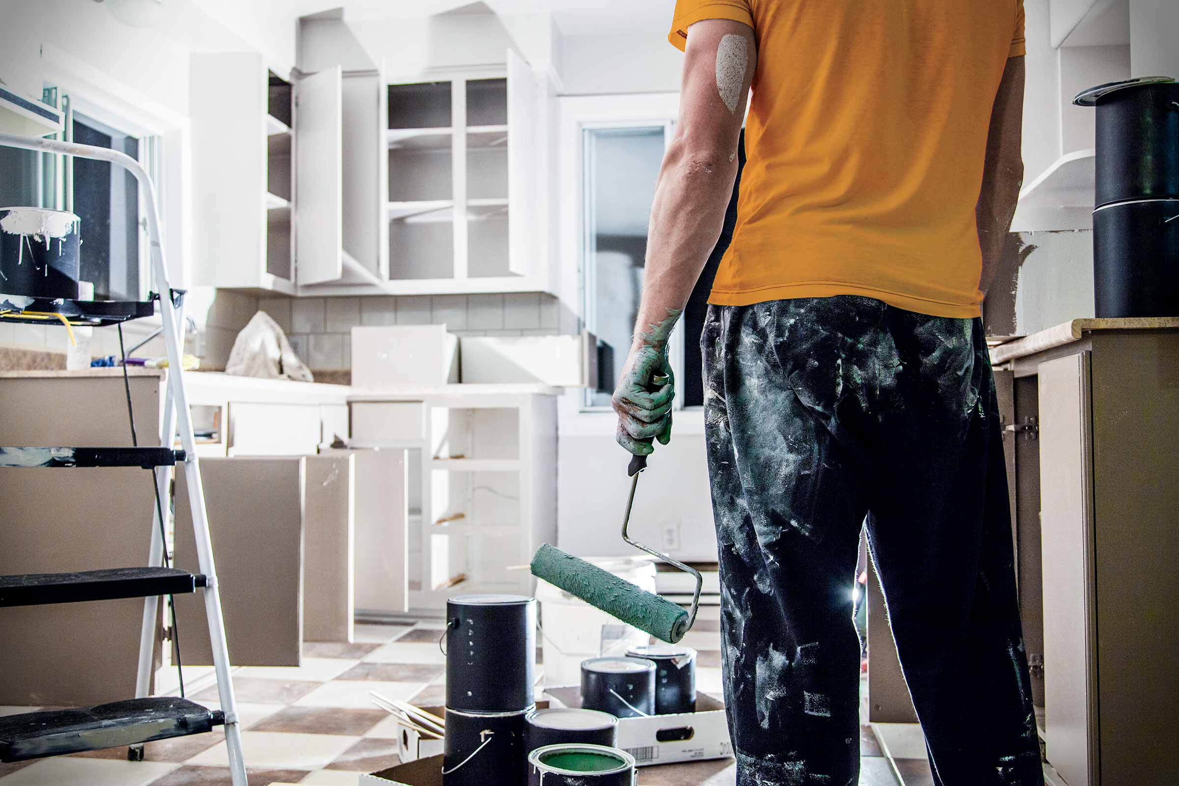 Interieurfoto van een gerenoveerde keuken met een schilder die een verfroller vasthoudt, verfemmers, een ladder en gedeeltelijk gemonteerde keukenkasten.