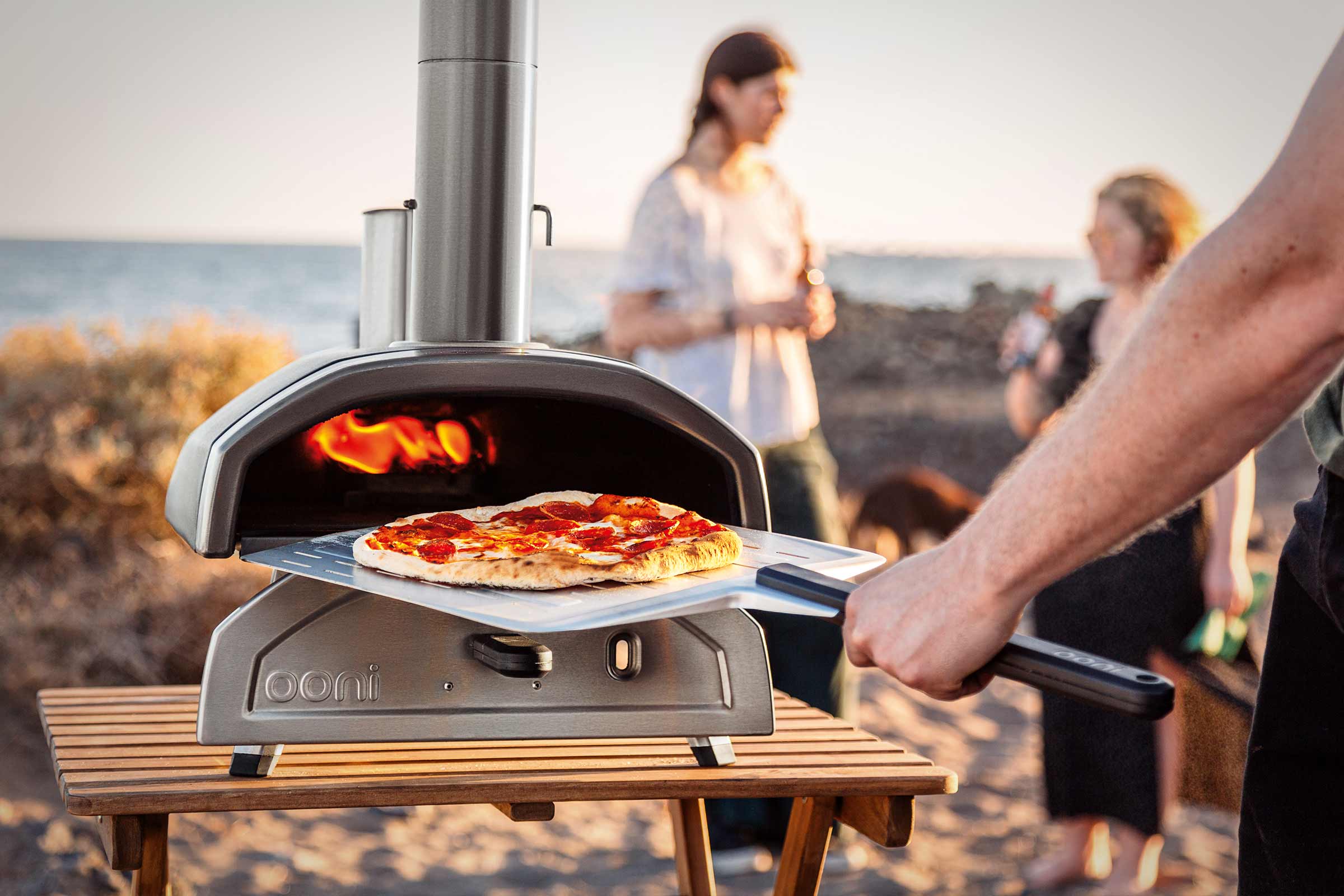 Ooni pizzaoven met pizzaschep op een houten tafel, met mensen op het strand op de achtergrond.